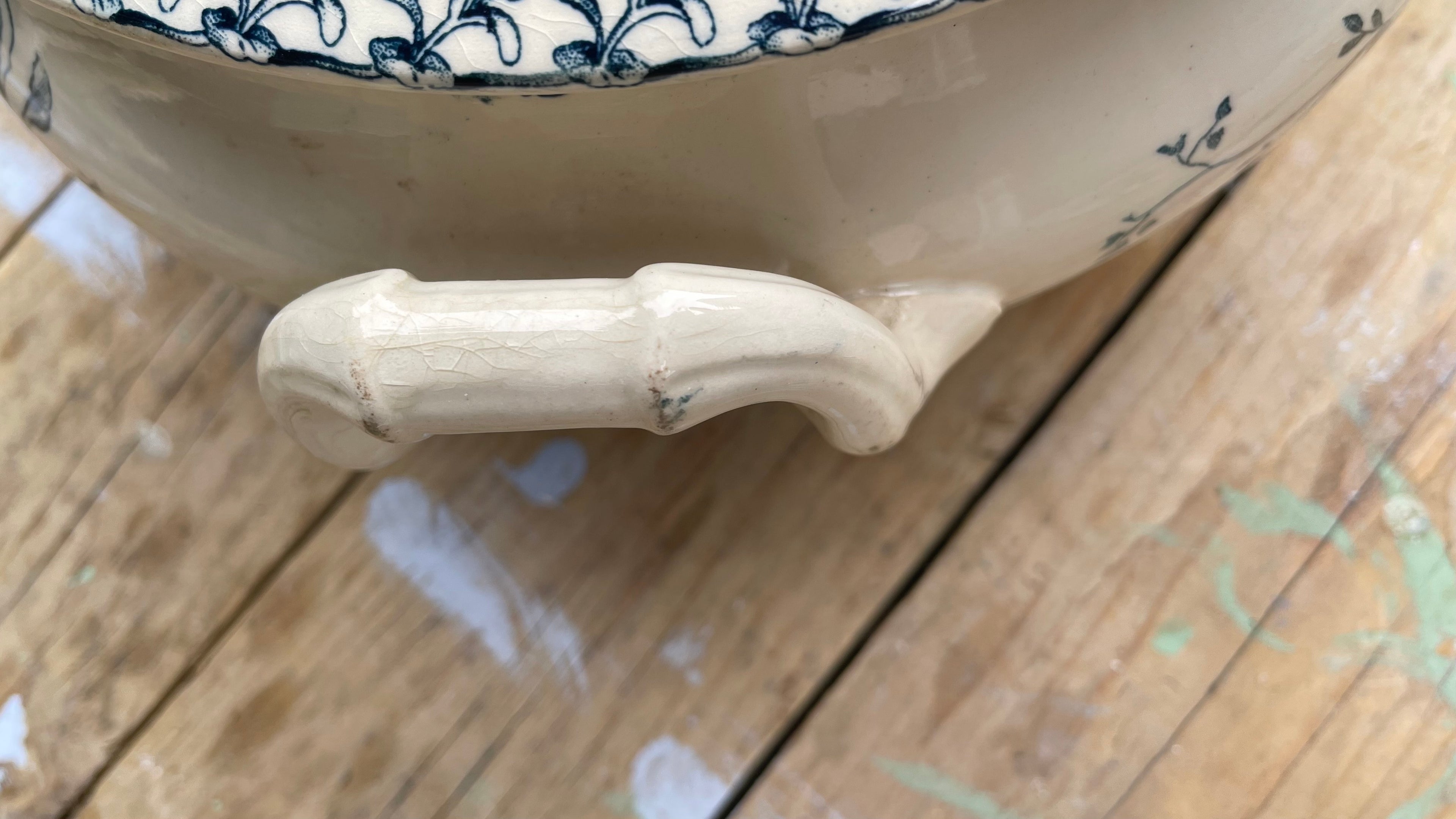 Close-up of a ceramic bowl or tureen with a glossy cream-colored handle and blue floral pattern on the rim, resting on a wooden surface.
Details