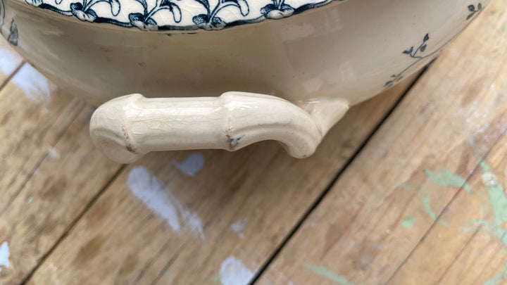 Close-up of a ceramic bowl or tureen with a glossy cream-colored handle and blue floral pattern on the rim, resting on a wooden surface.
Details