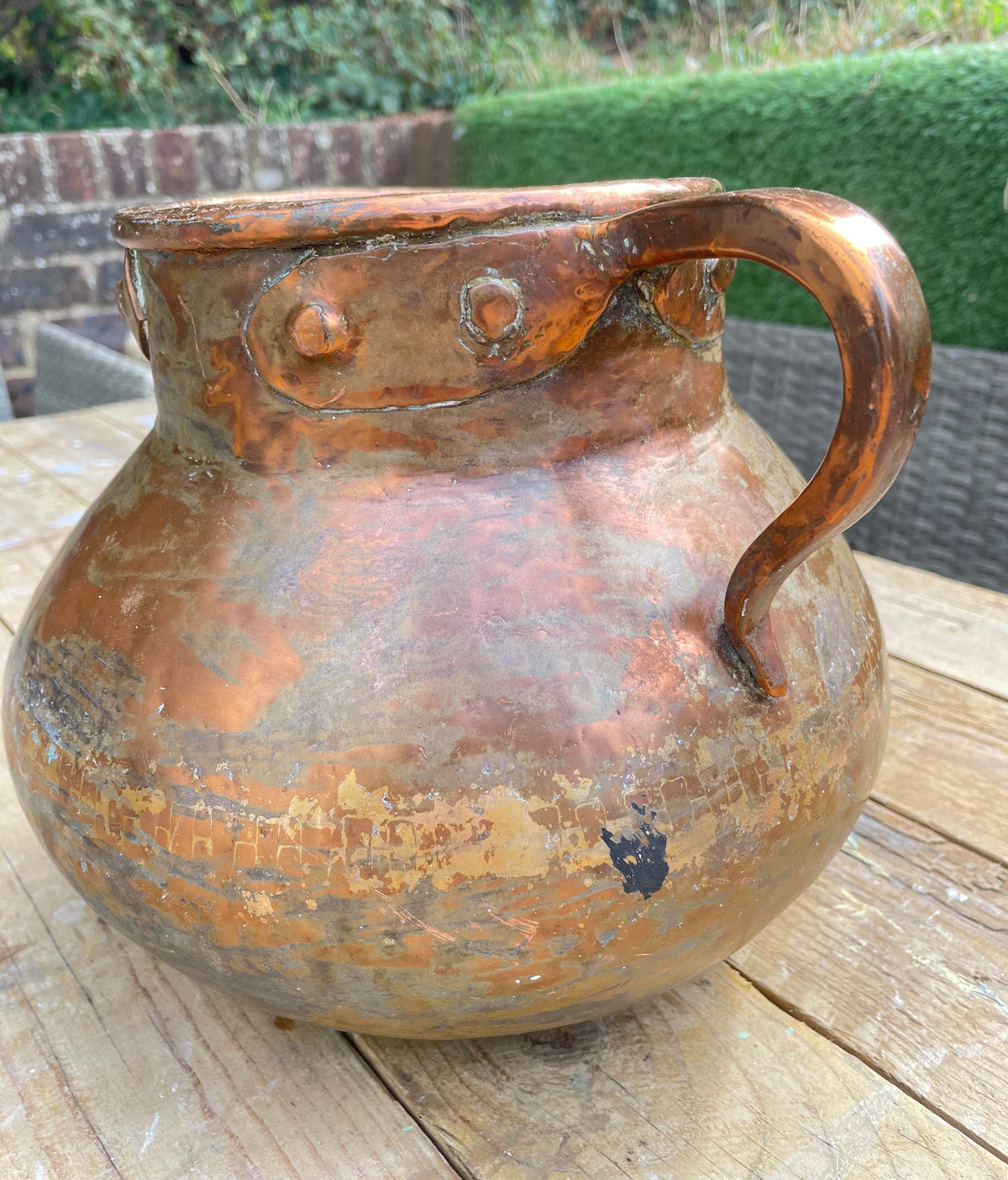 rustic copper-colored metal pitcher with a rounded body, curved handle, and riveted rim, photographed on a wooden table with an outdoor garden background.
