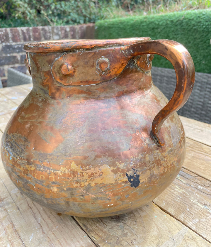 rustic copper-colored metal pitcher with a rounded body, curved handle, and riveted rim, photographed on a wooden table with an outdoor garden background.
