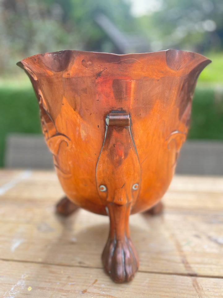 A copper-colored metal bucket or planter with a flared rim, decorative embossed scrollwork, and a central handle, shown standing on a wooden surface with a blurred outdoor background.
