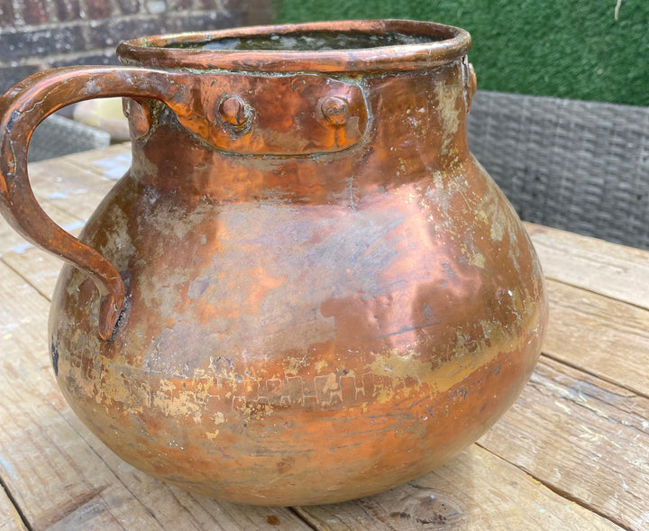 A rustic copper-colored metal pitcher with a curved handle and rivet details, sitting on a wooden table with an outdoor garden background.