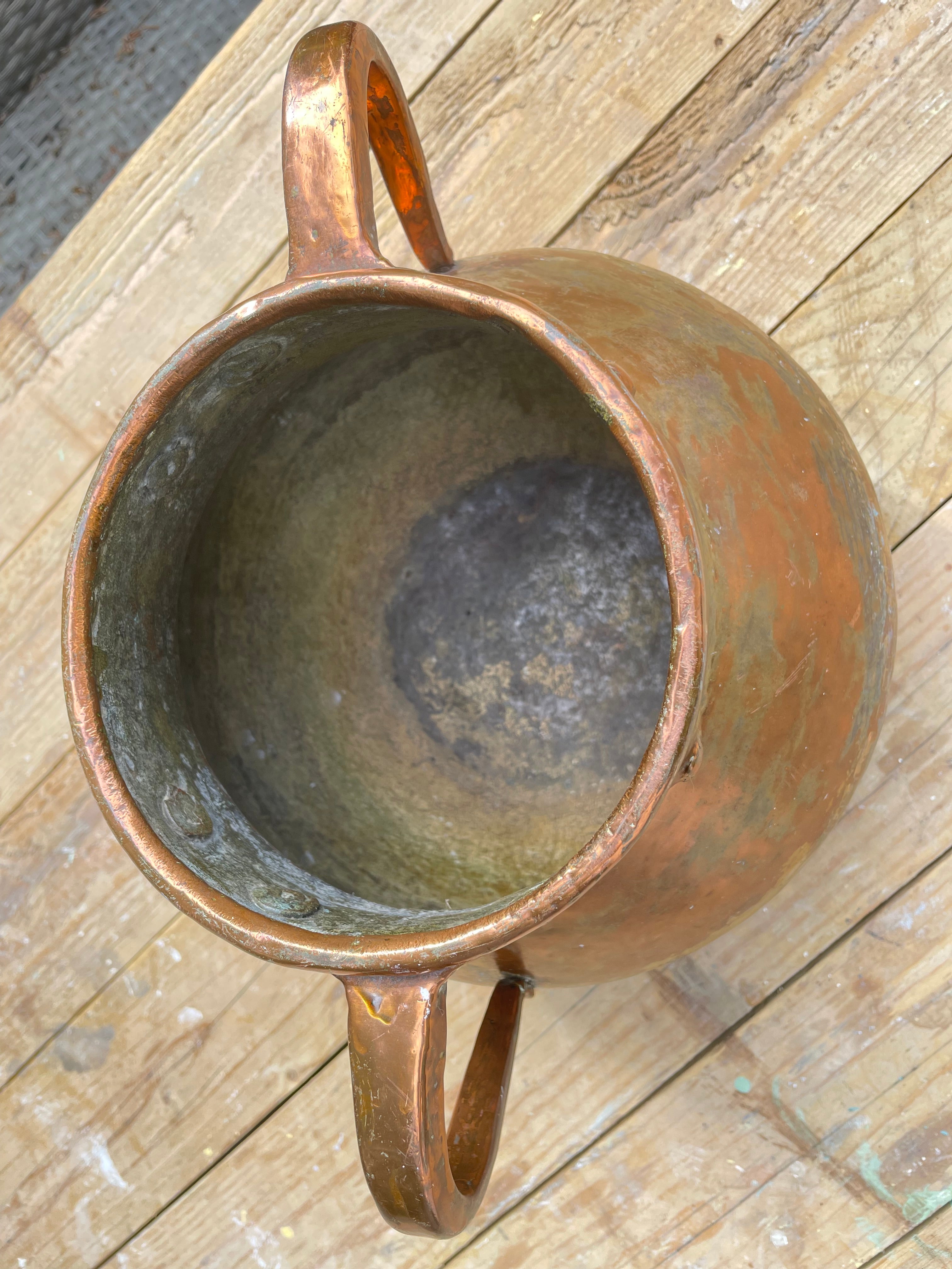 A copper-colored metal pitcher with two loop handles, photographed from above on a rustic wooden plank surface.
Details