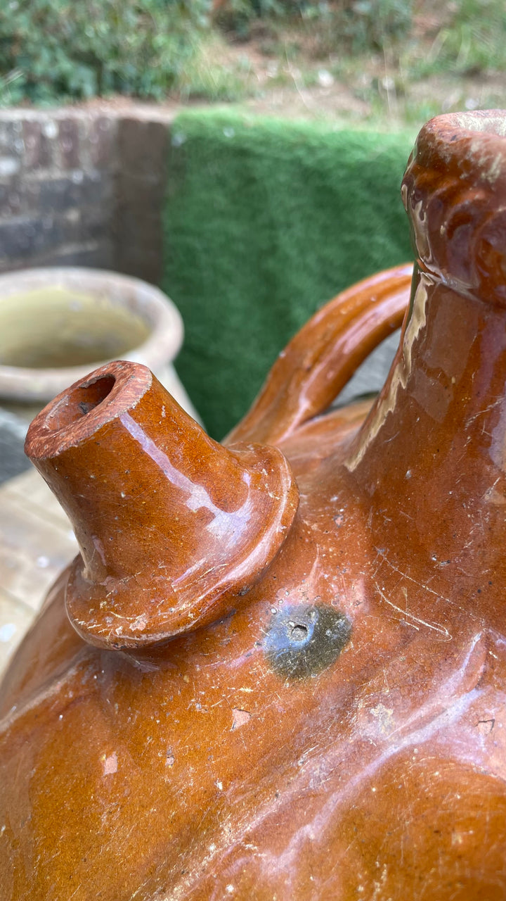 Close-up of a rustic terracotta clay jug or pitcher with a glossy brown-orange glaze, showing the spout and shoulder against an outdoor background.
