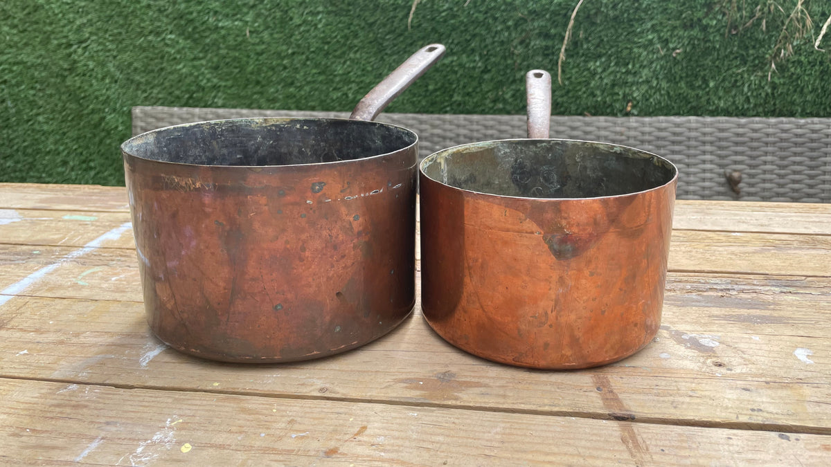 Two vintage copper saucepans with long handles sitting on a wooden table outdoors against a green hedge background.