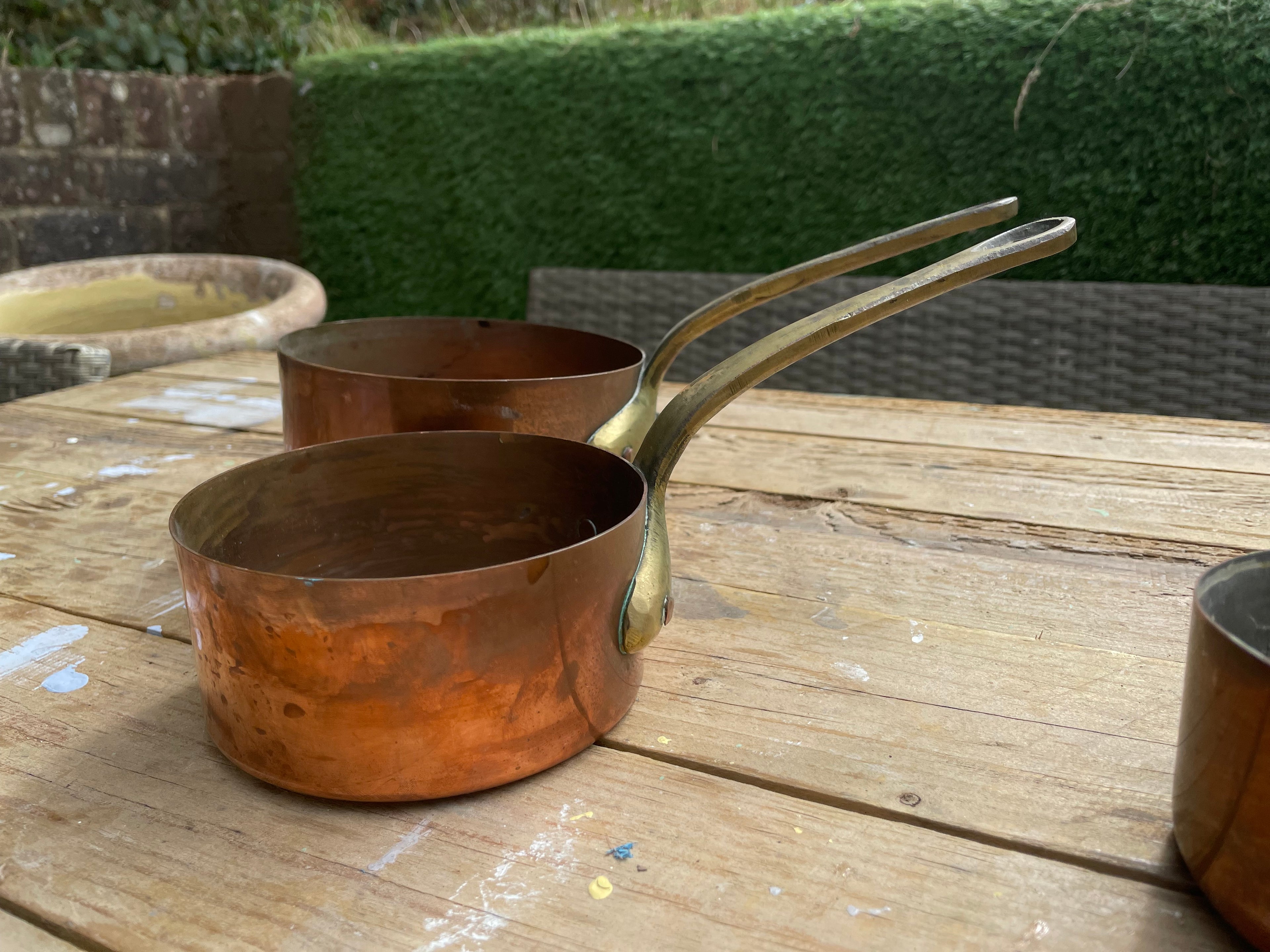 Two copper saucepans with long handles sitting on a wooden table outdoors, with a hedge and patio furniture in the background.
Details