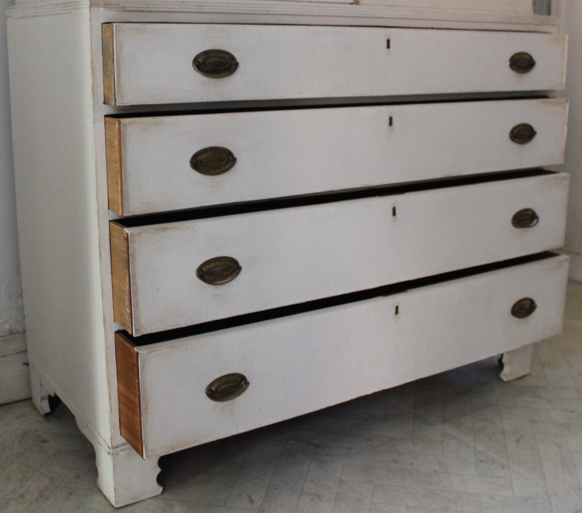 A vintage white painted wooden chest of drawers with four drawers and dark oval handles, set in a room with a tiled floor and white walls.
