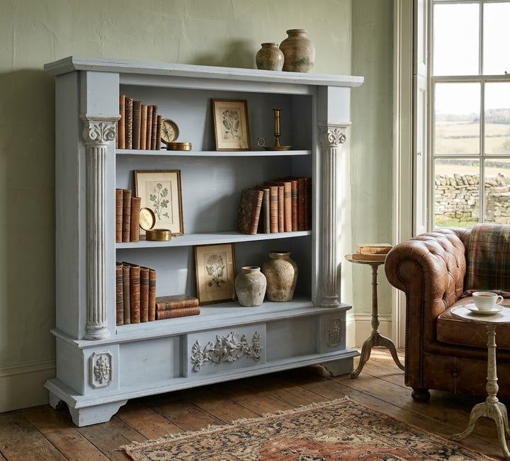 A tall, light blue-gray wooden bookcase with decorative columns and carved details, styled with books and vases in a rustic room next to a brown leather armchair.