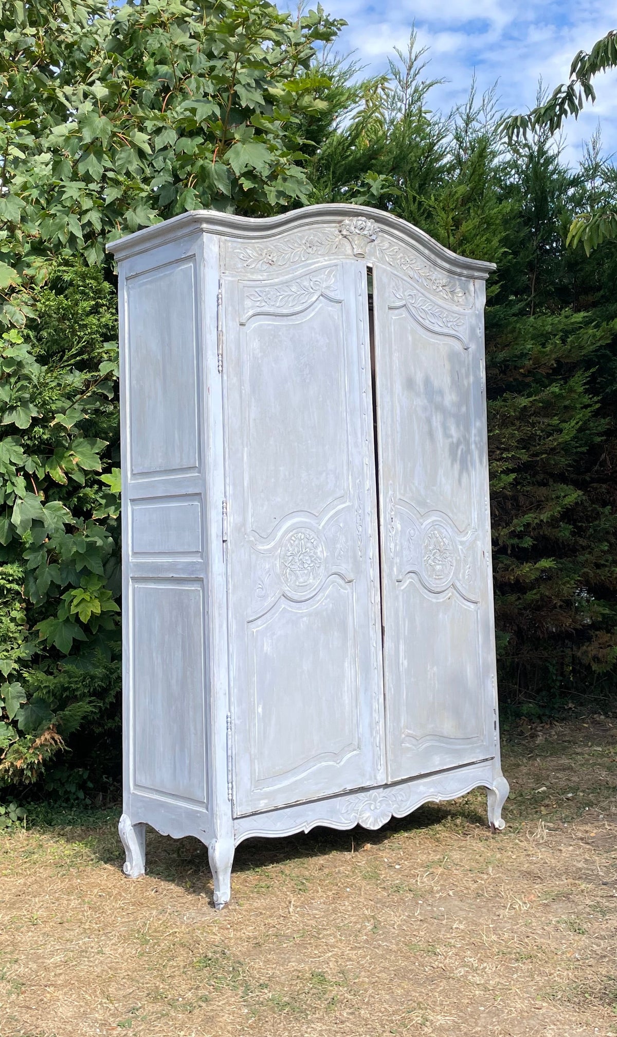 A distressed white painted wooden three-door wardrobe or armoire cabinet, photographed outdoors on a grassy lawn with trees and blue sky in the background.