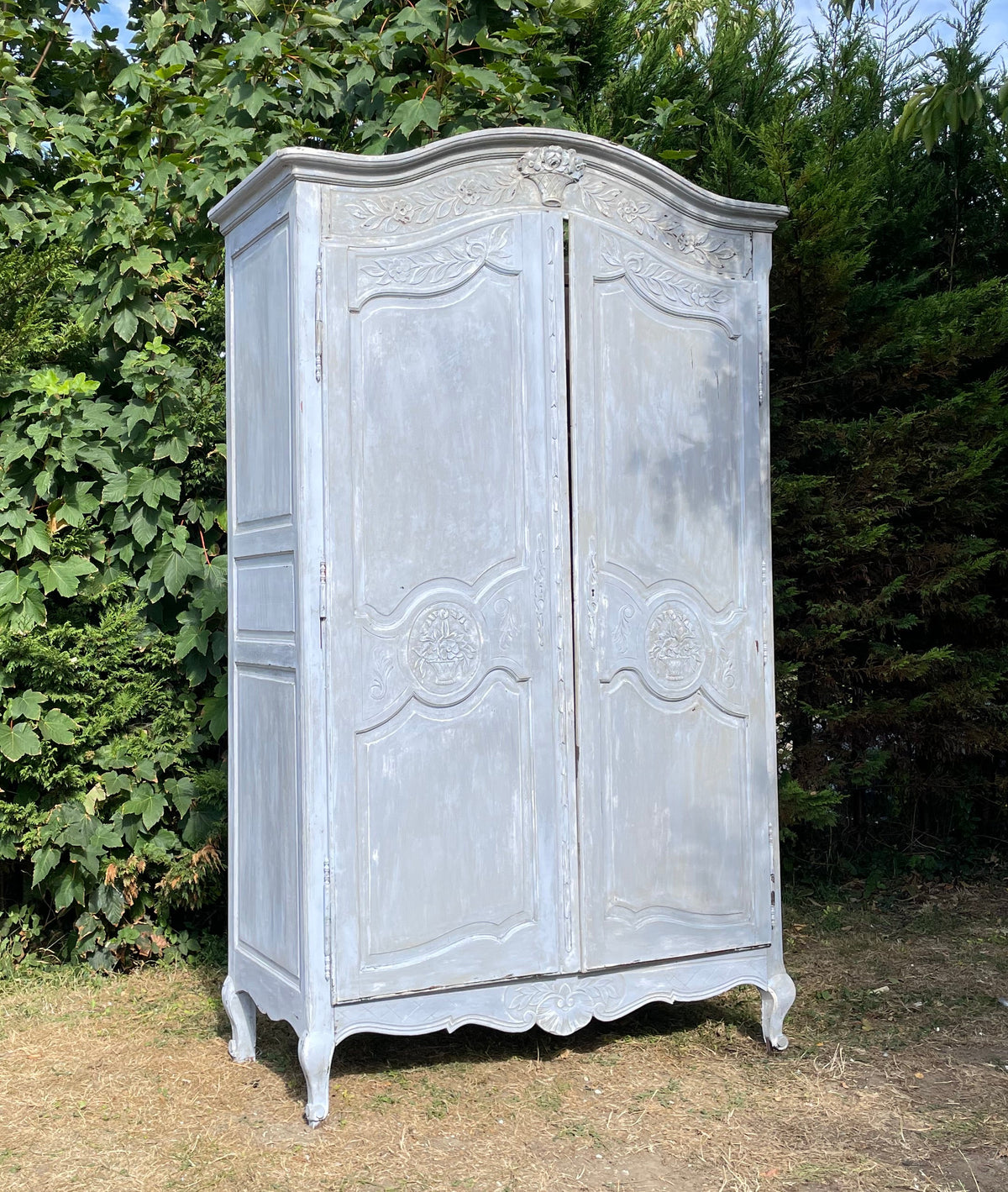 A distressed light gray painted wooden cabinet or wardrobe photographed outdoors on grass with trees and blue sky in the background.