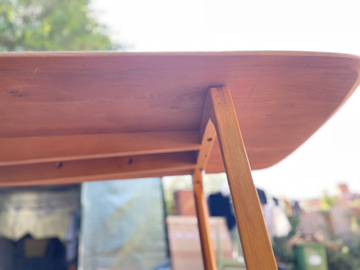 Close-up of a wooden table top and leg in warm medium brown wood, photographed outdoors with a blurred background.