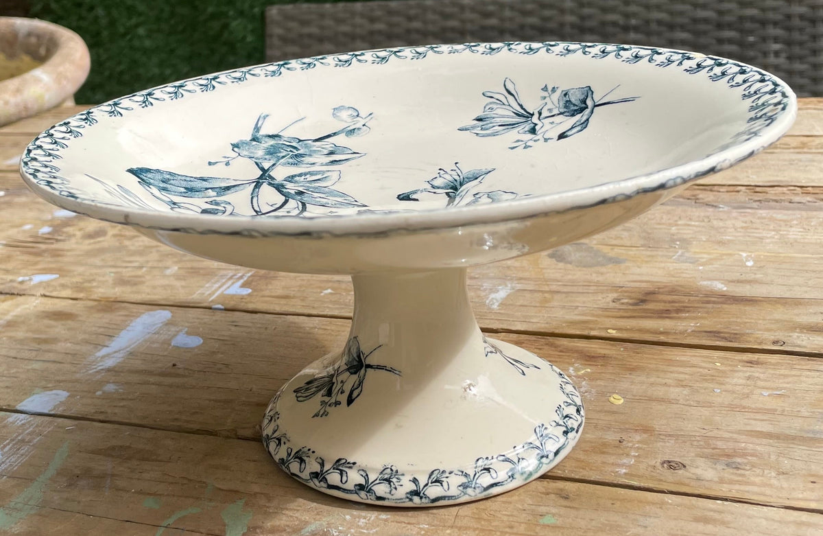 A ceramic pedestal plate with a blue floral and butterfly pattern on an off-white background, set on a wooden table outdoors.