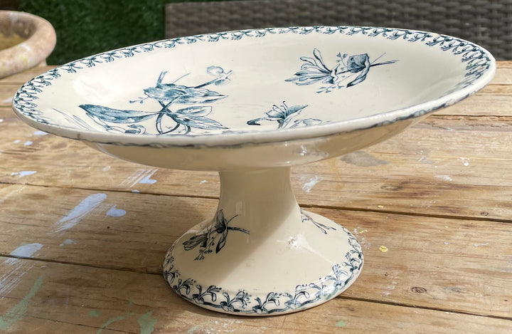 A ceramic pedestal plate with a blue floral and butterfly pattern on an off-white background, set on a wooden table outdoors.