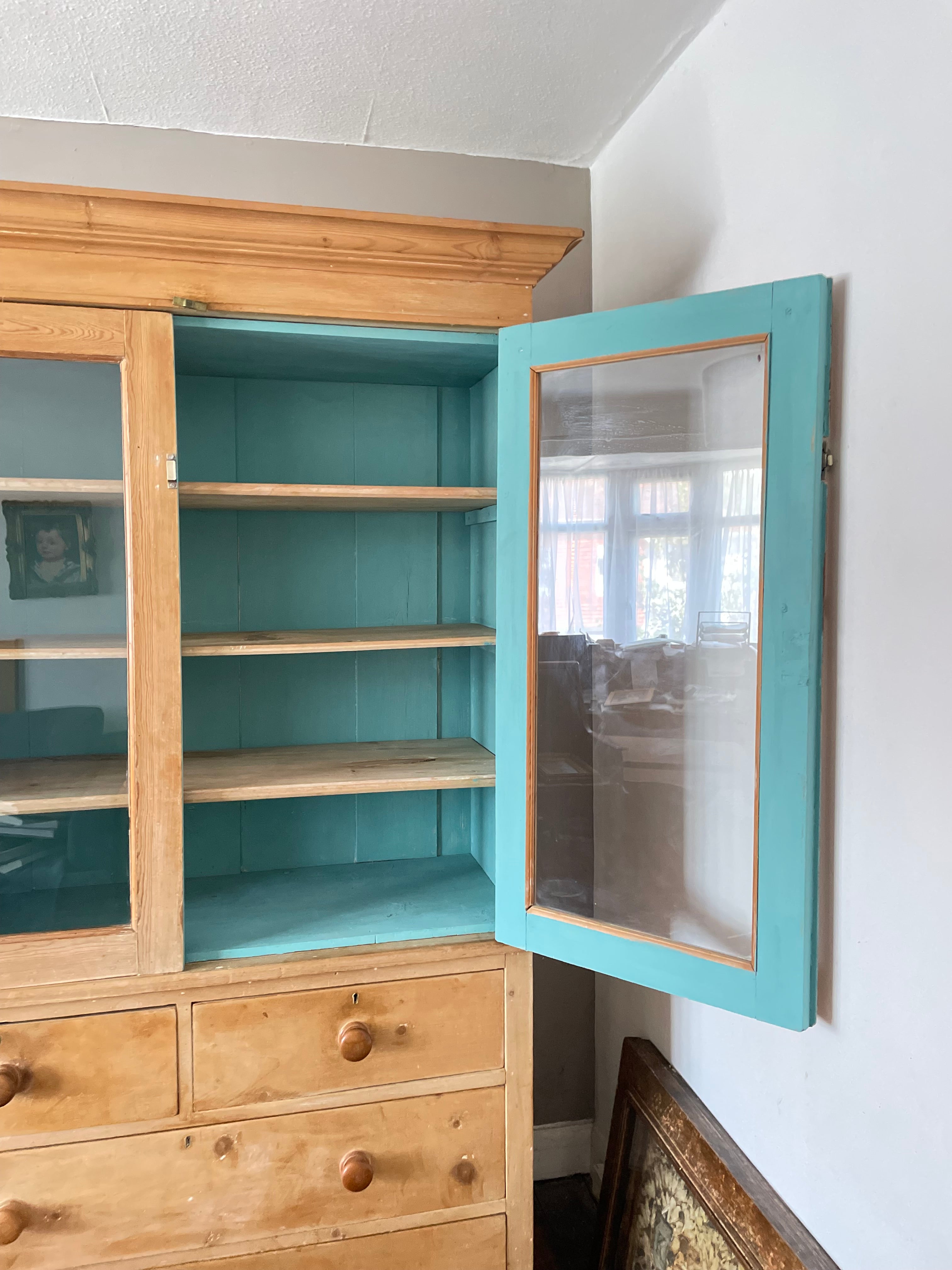 A wooden cabinet with turquoise painted interior shelves and a matching turquoise glass-front door, shown open against a white wall.