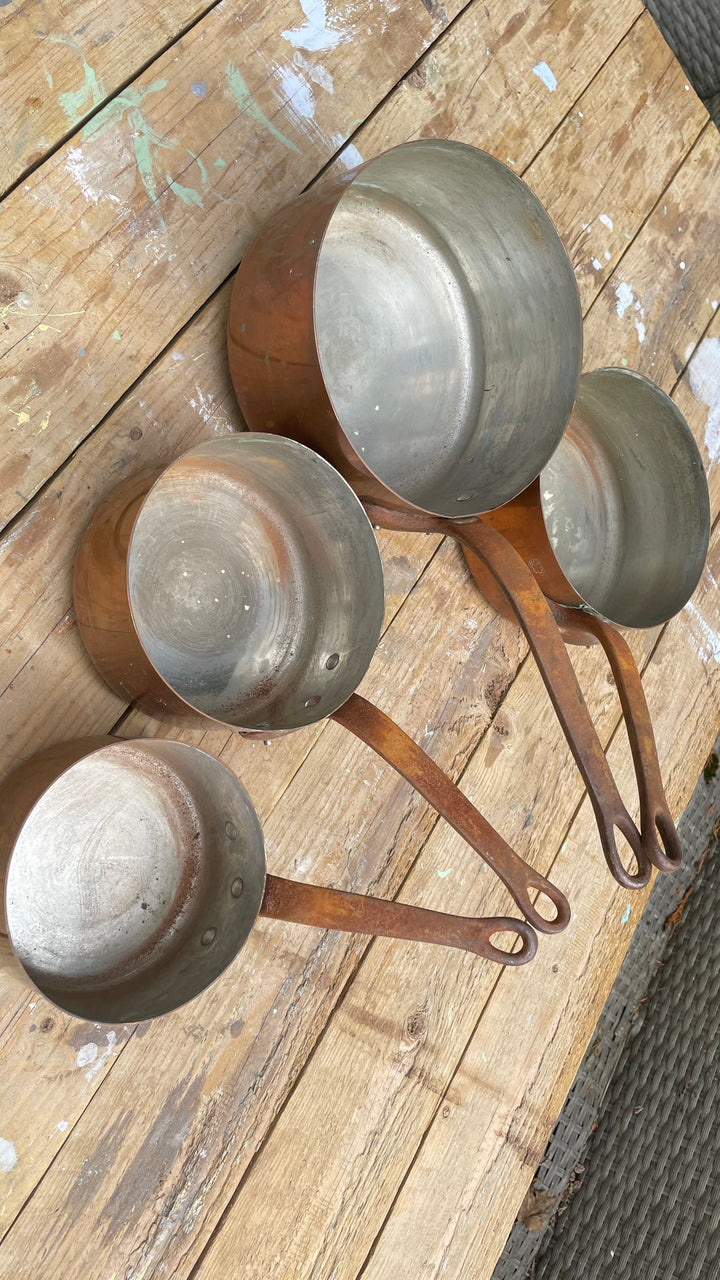  vintage copper and stainless steel measuring cups with long handles, arranged on a rustic wooden plank surface.
