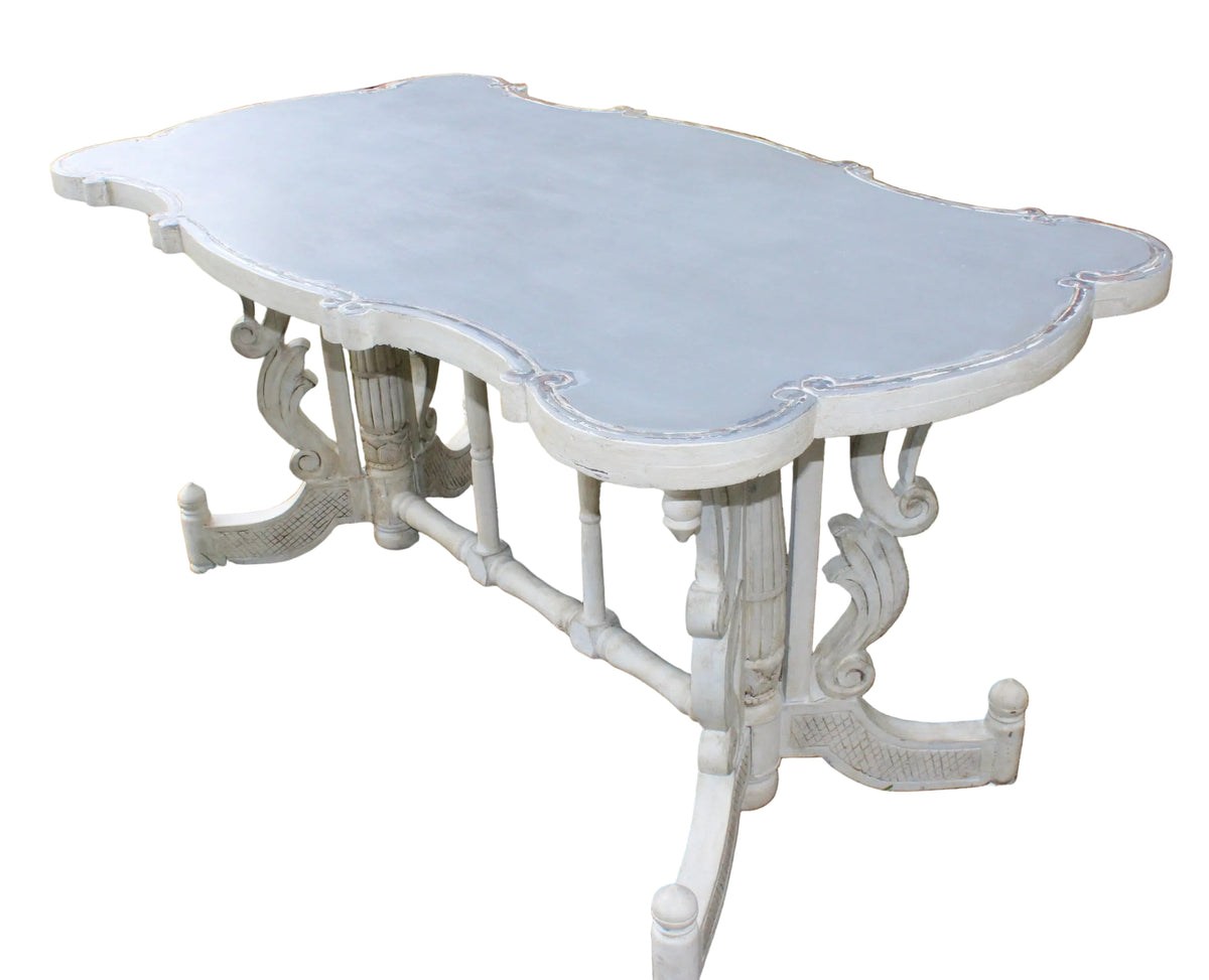 A large, ornate outdoor dining table with a scalloped slate-blue top and carved white legs