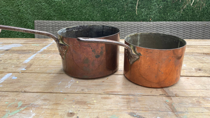 Two copper saucepans, one larger and one smaller, resting on a wooden table with a green hedge in the background.