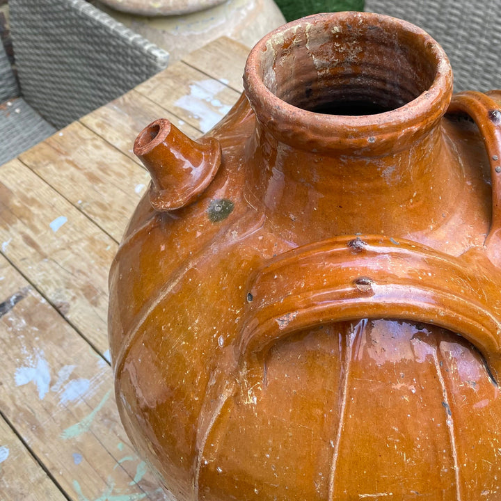 large rustic terracotta clay vase with a spout and ribbed body, photographed on a wooden table in an outdoor patio setting with a smaller pot and woven chair visible in the background.