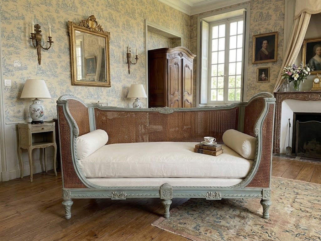 vintage French-style chaise lounge with a pale blue-green painted frame, woven cane backrest, and cream cushions, set in an elegant, classic room with patterned wallpaper, a fireplace, and wooden floors.