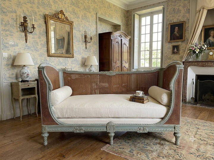 vintage French-style chaise lounge with a pale blue-green painted frame, woven cane backrest, and cream cushions, set in an elegant, classic room with patterned wallpaper, a fireplace, and wooden floors.