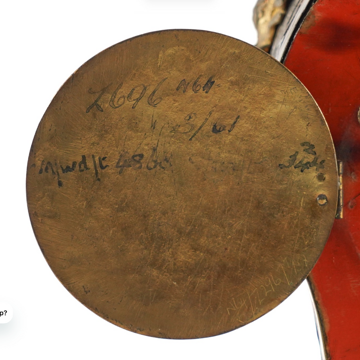 A close-up of a round, antique bronze-colored metal medallion or coin lid with handwritten markings, shown within a screenshot interface.