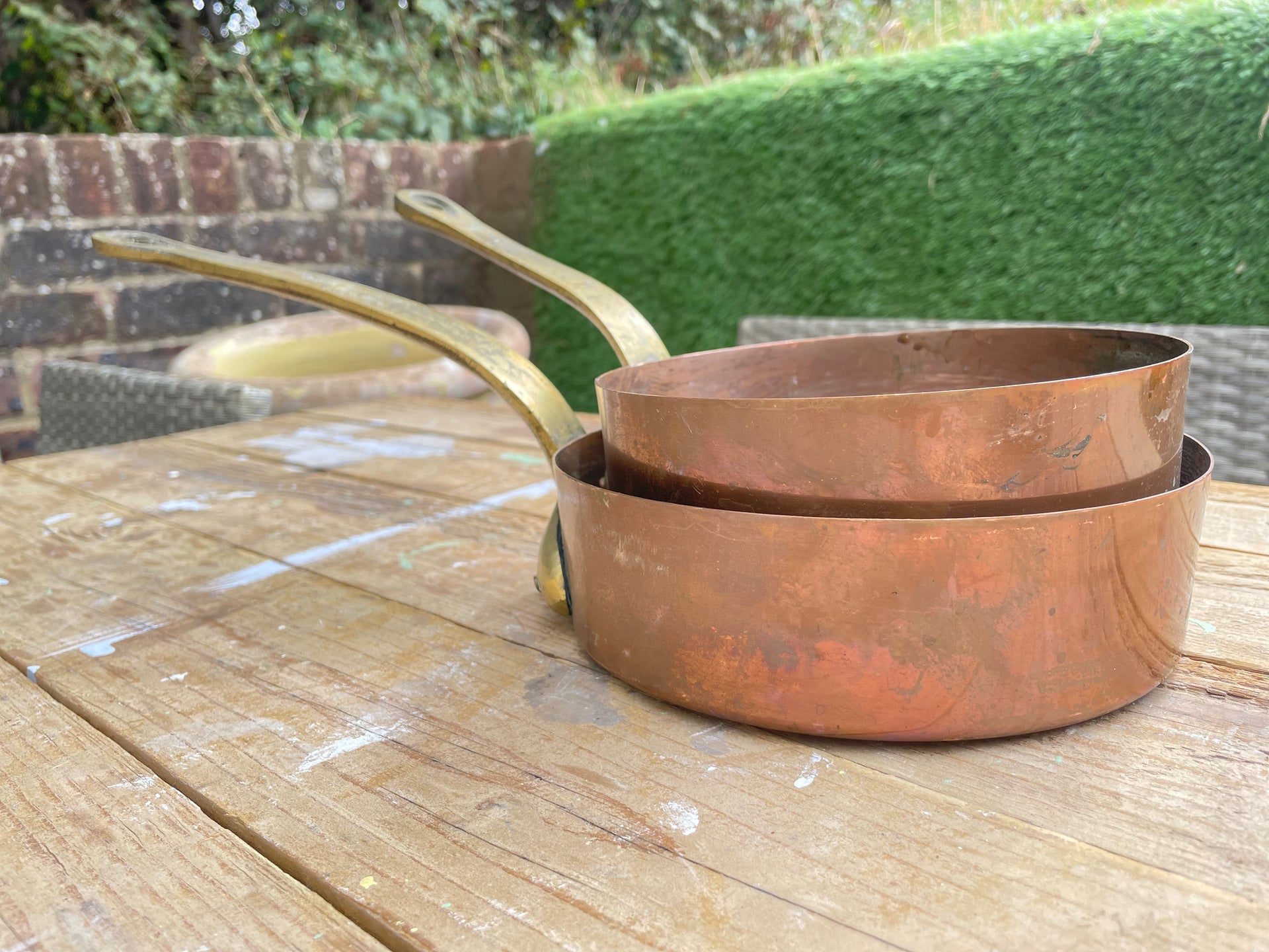  nested copper saucepans with long handles resting on a wooden table outdoors, with a hedge and brick wall in the background.
