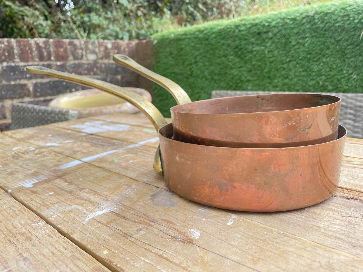  nested copper saucepans with long handles resting on a wooden table outdoors, with a hedge and brick wall in the background.
