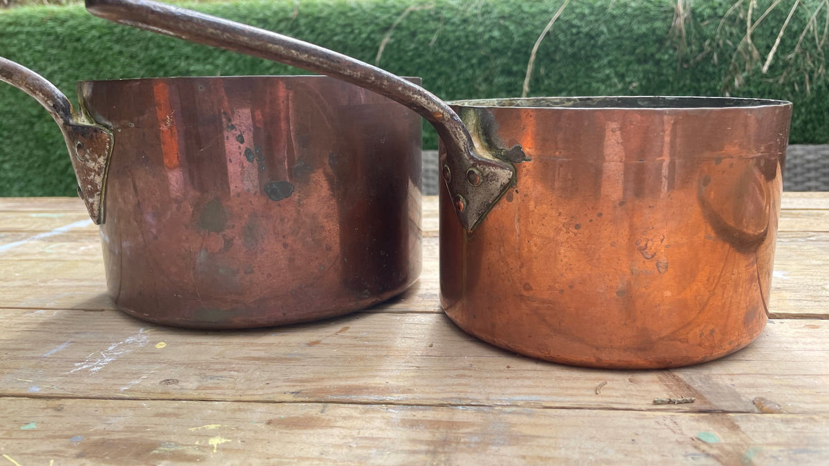 Two copper saucepans with long handles resting on a wooden table outdoors, with a green hedge in the background.