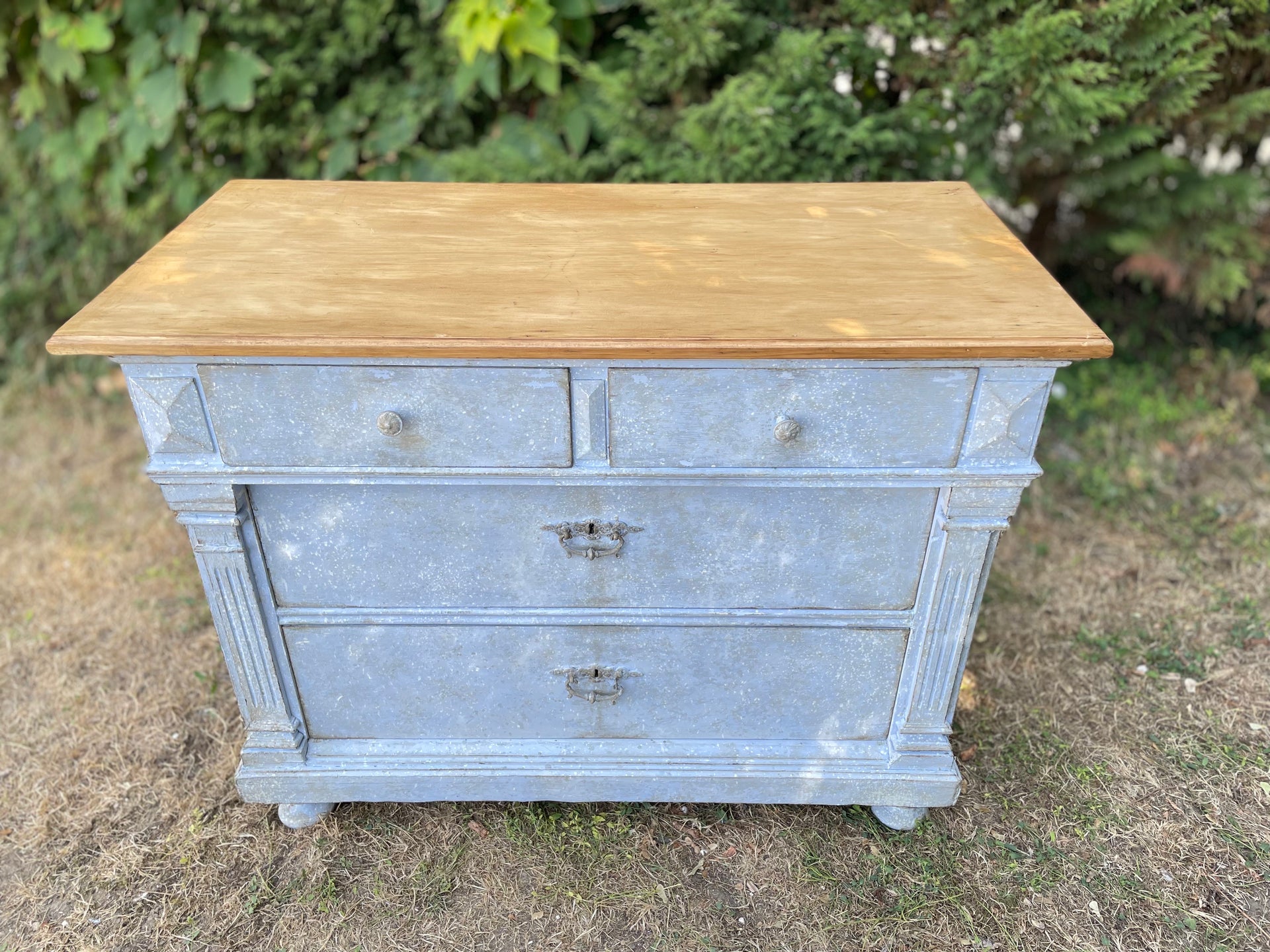 A vintage-style three-drawer chest or dresser with a distressed light blue-gray painted body and a natural light brown wood top, photographed outdoors on grass with green foliage in the background.