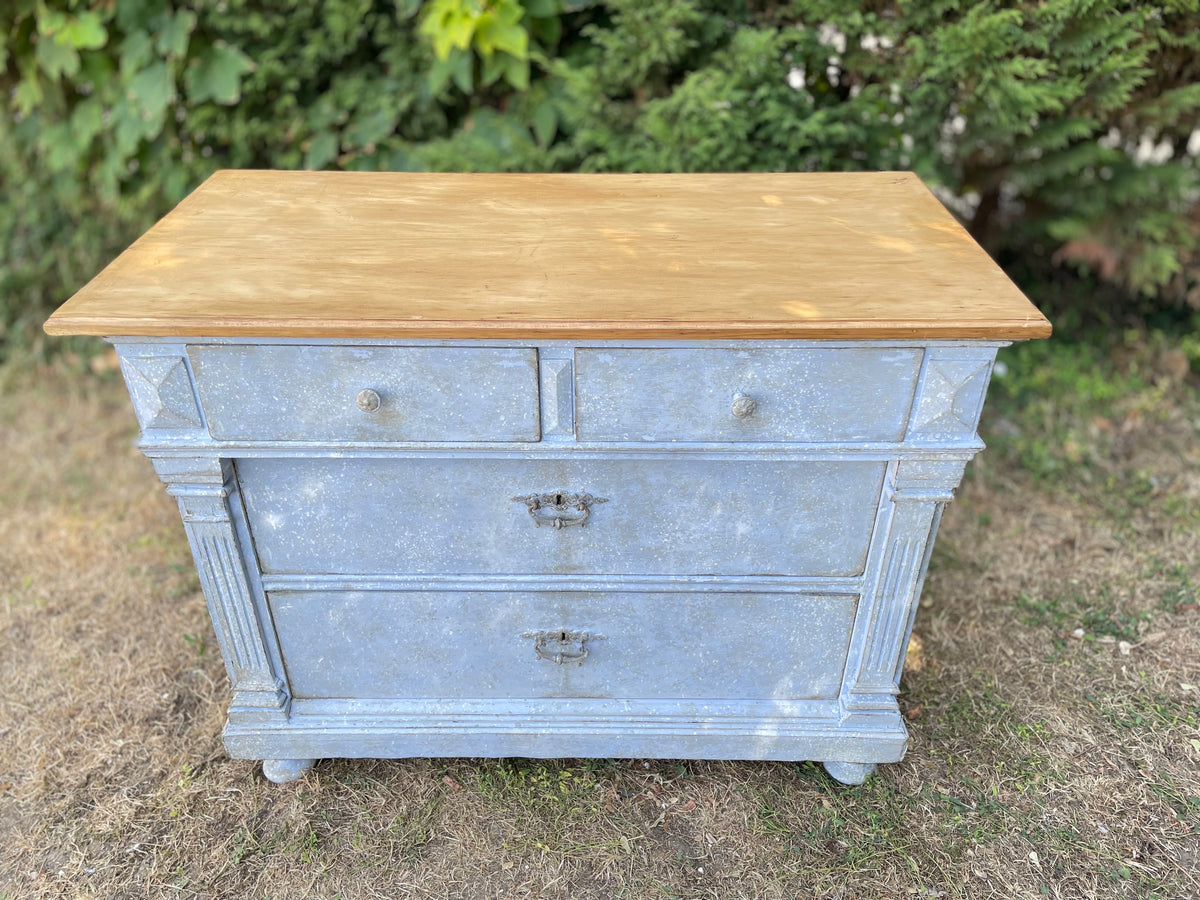 A vintage-style three-drawer chest or dresser with a distressed light blue-gray painted body and a natural light brown wood top, photographed outdoors on grass with green foliage in the background.