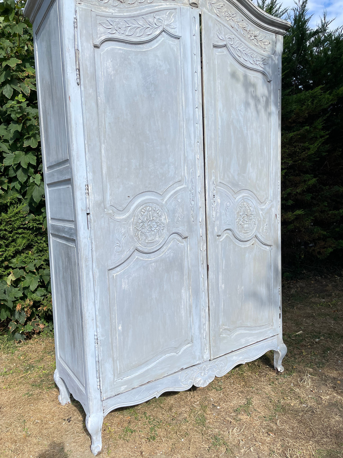 A large, vintage-style painted wooden cabinet or armoire with ornate carved details and a distressed light gray finish, photographed outdoors on grass and foliage.