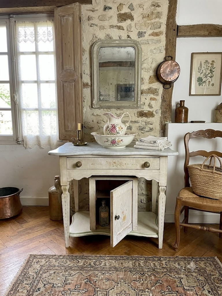 An Antique painted white distressed table with a marble top, set in a rustic room with a stone wall, wooden door, and pattern