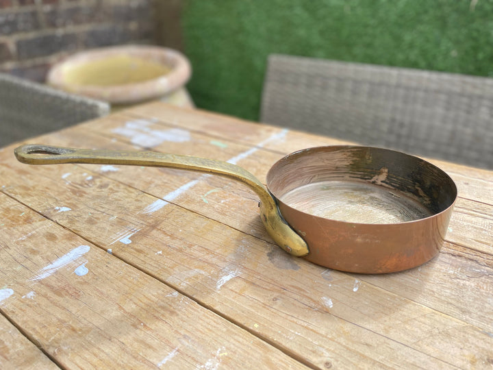 small copper saucepan with a long handle, resting on a weathered wooden table outdoors.