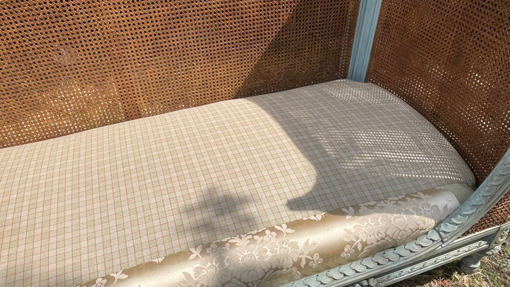 A close-up of a woven cane daybed or lounger with a beige and white checkered cushion and a cream floral-patterned bolster, set against a brown woven cane backrest.