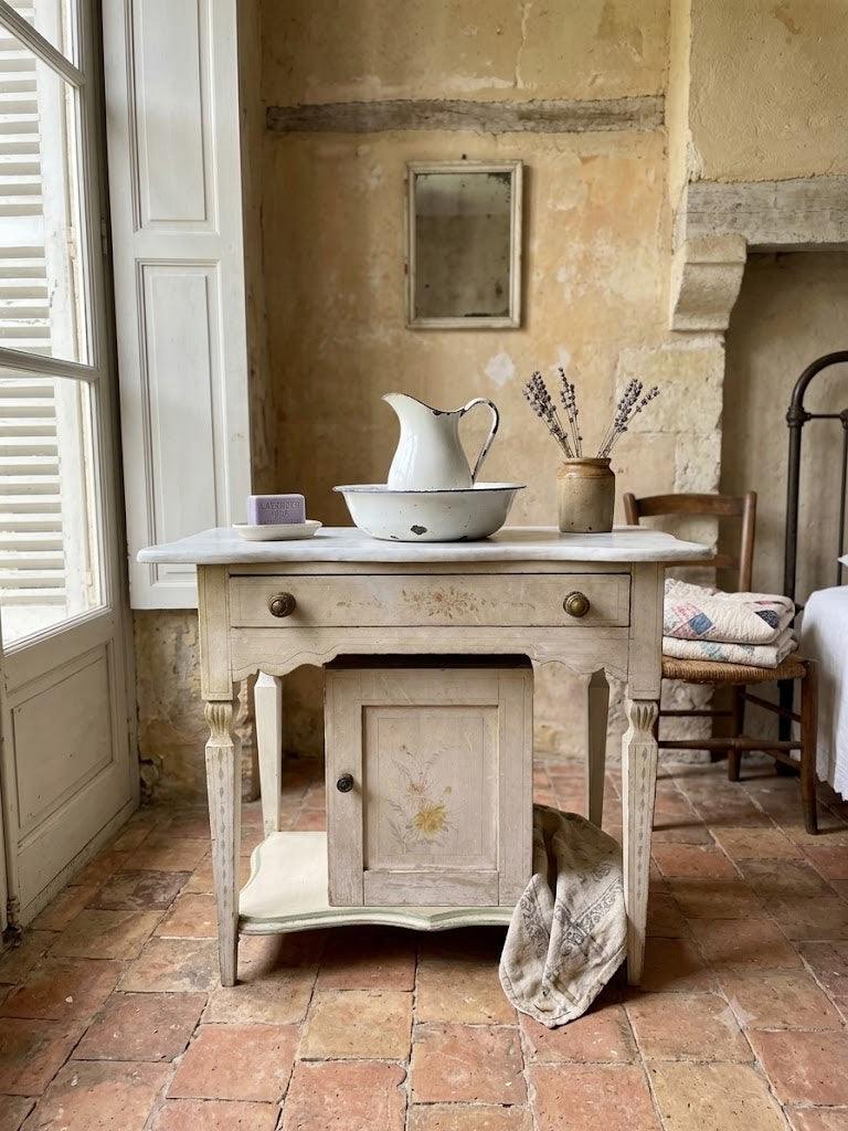 A distressed, vintage-style French provincial side table with a whitewashed finish and brass knobs, styled in a rustic room with terracotta tile floors and a stone wall.