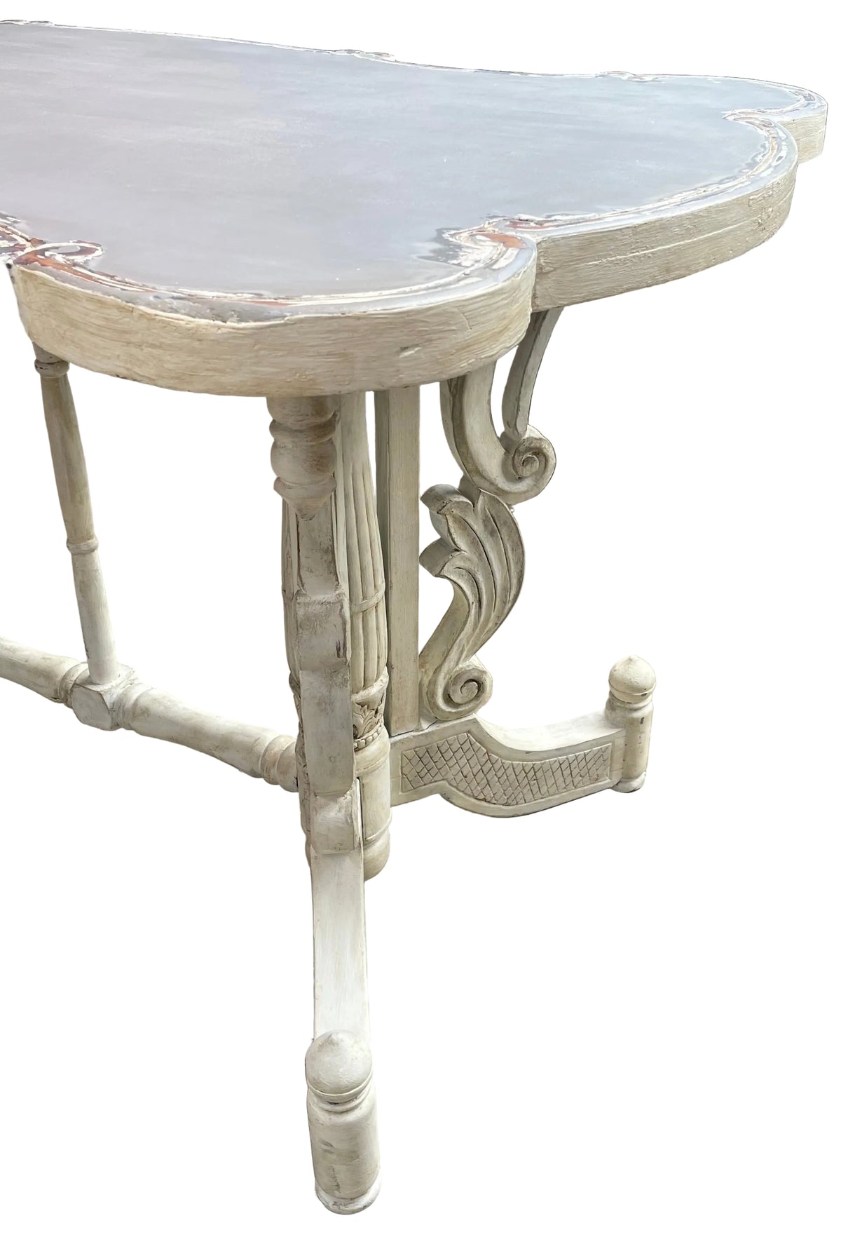 A distressed white carved dining table with a gray top, photographed outdoors on a tiled patio with a green hedge and wicker furniture in the background.