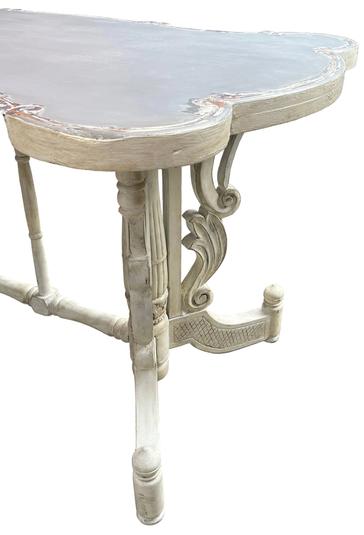 A distressed white carved dining table with a gray top, photographed outdoors on a tiled patio with a green hedge and wicker furniture in the background.