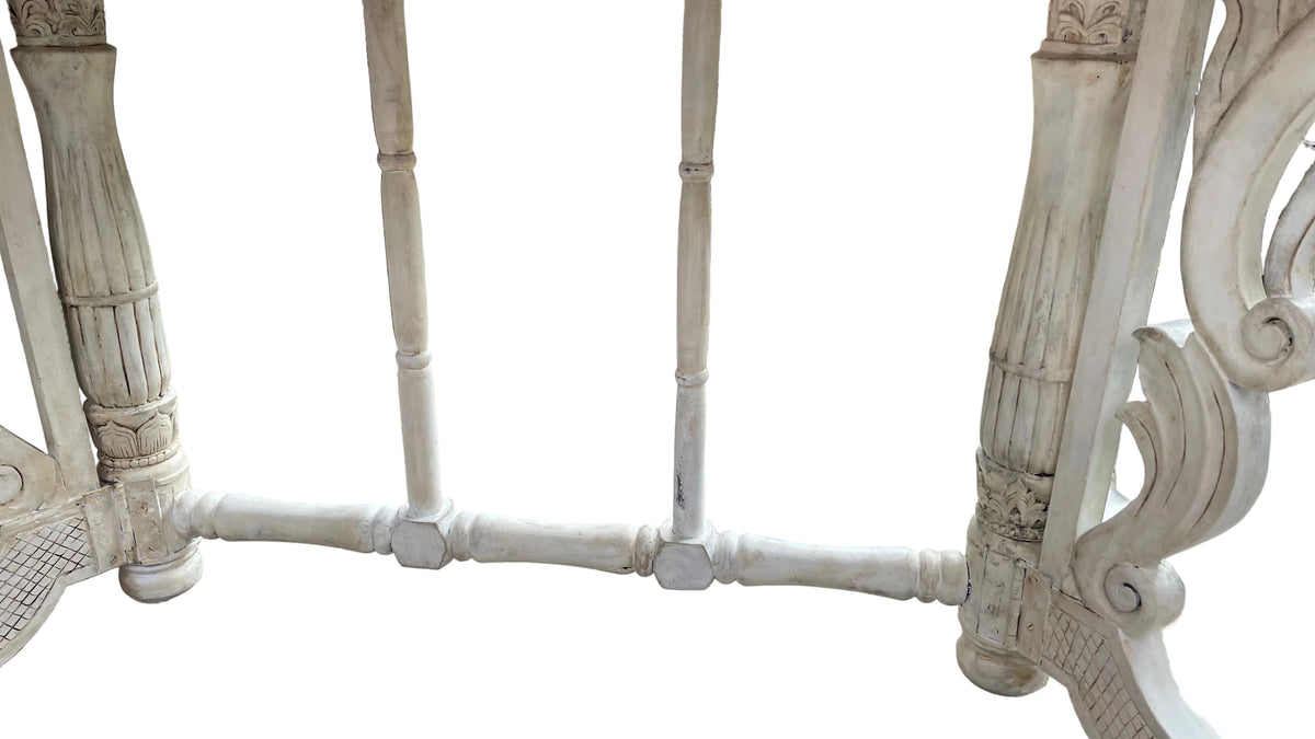 close-up of an ornate white carved wooden or metal frame structure, possibly a table base or decorative stand, with vertical spindles and scrollwork details, set on a tiled patio with a wicker sofa in the background.