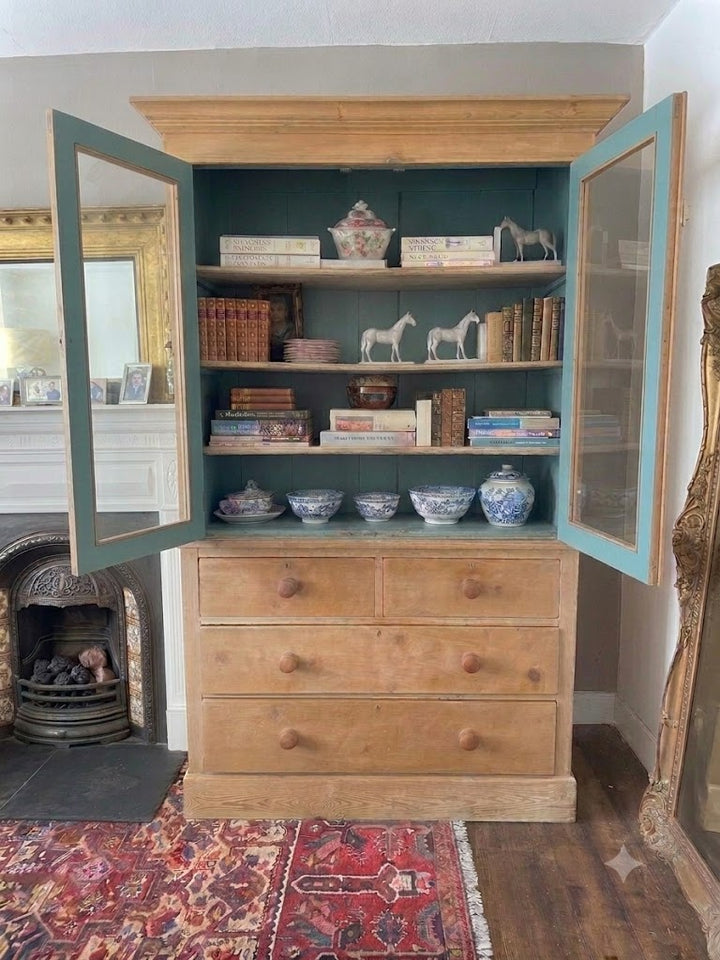 A large wooden cabinet with two open doors, painted teal on the interior and drawers, filled with books and decorative ceramic vessels, set in a room with a fireplace and patterned rug.