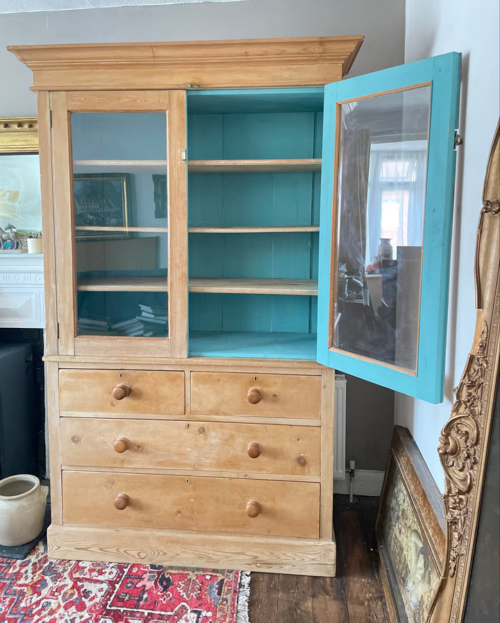 A vintage wooden cabinet with a turquoise painted upper frame and glass doors, shown next to a light natural wood chest of drawers with round knobs in an indoor room setting.
