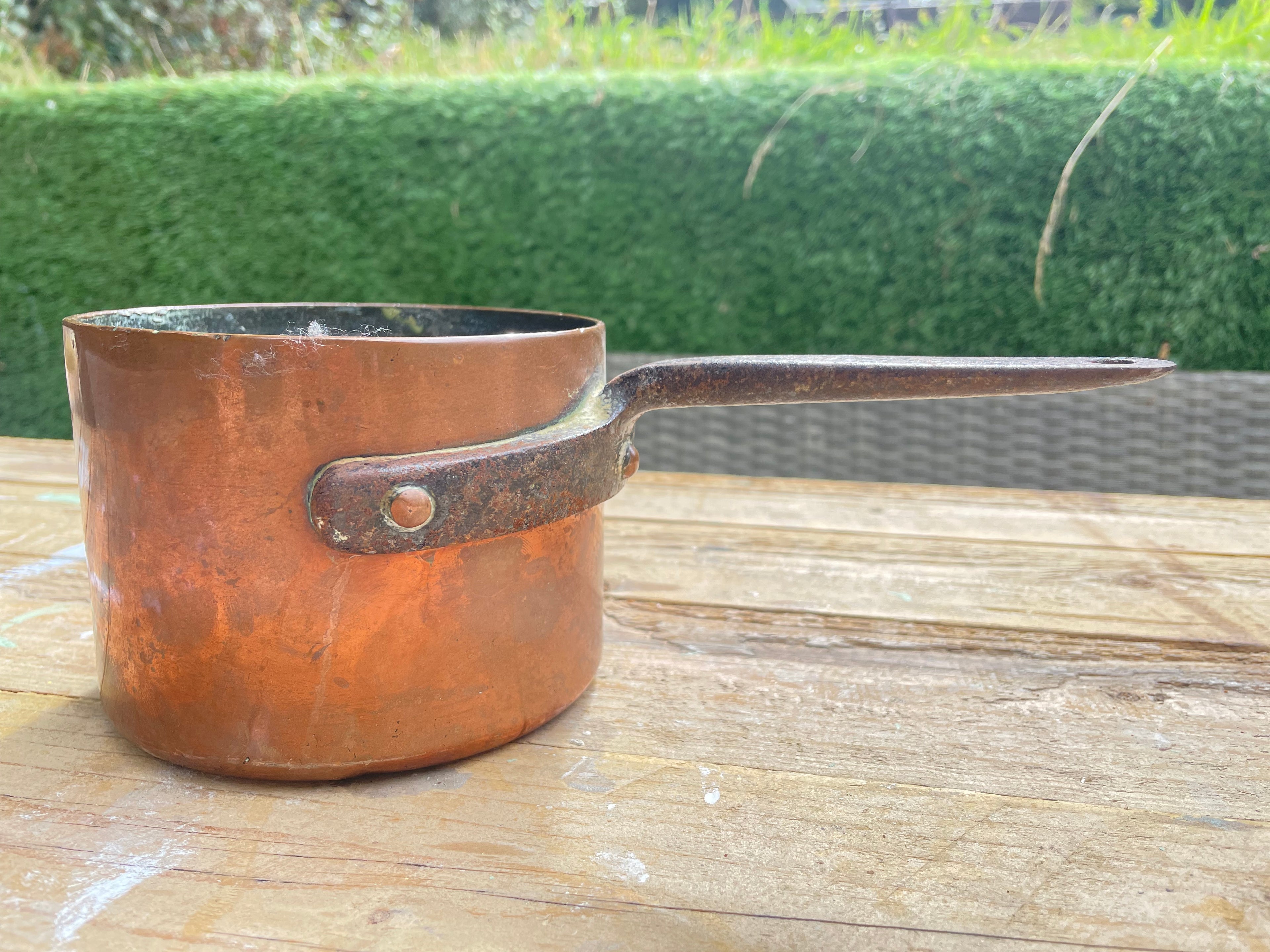 A vintage copper saucepan with a long dark metal handle, resting on a wooden table with a green hedge in the background.