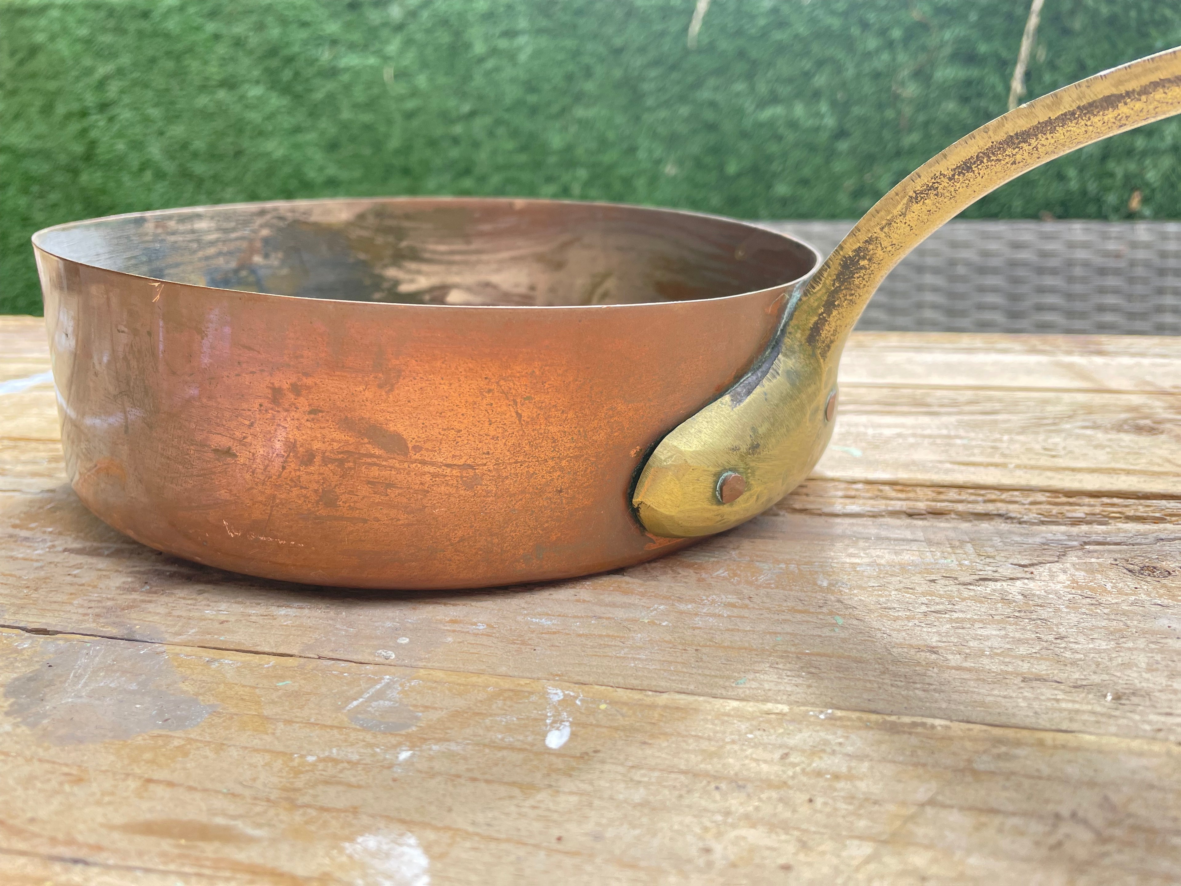 copper saucepan with a brass-colored handle and rivets, resting on a wooden surface with a green hedge in the background. Close Up