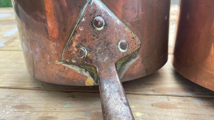 Close-up of a copper-colored metal pot or canister with a rusted triangular metal bracket and rivets, resting on a wooden surface.
