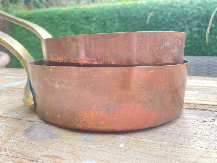 Two nested copper saucepans with brass handles resting on a wooden surface, with a blurred green hedge in the background