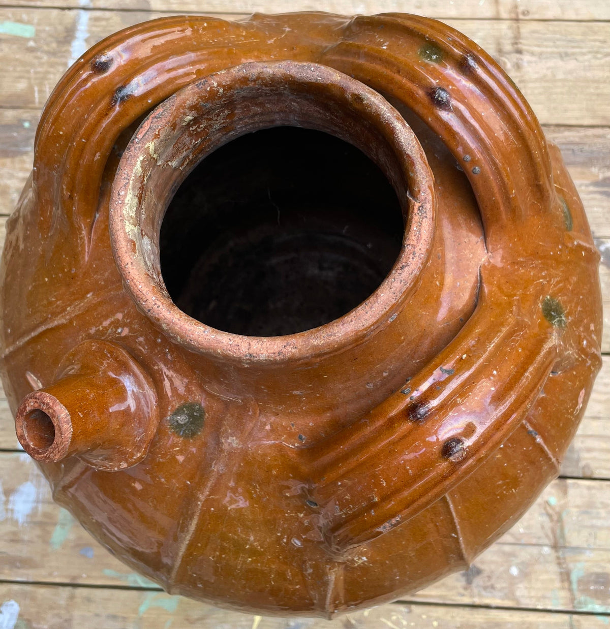 A rustic brown glazed ceramic pot with a small spout, photographed from above on a worn wooden surface.