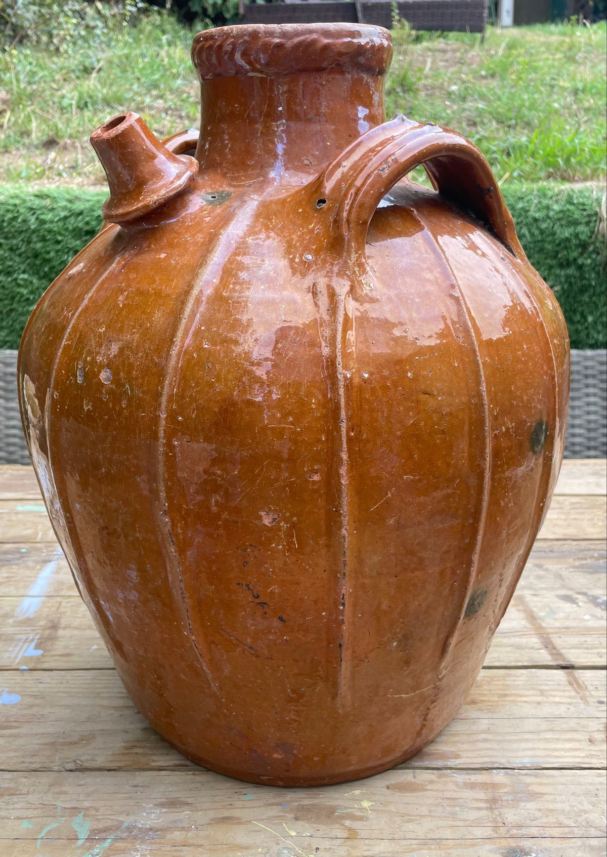 A large glazed ceramic amphora-style jug with a rounded body, shoulder spout, and side handle, photographed outdoors on a wooden surface with garden greenery in the background.