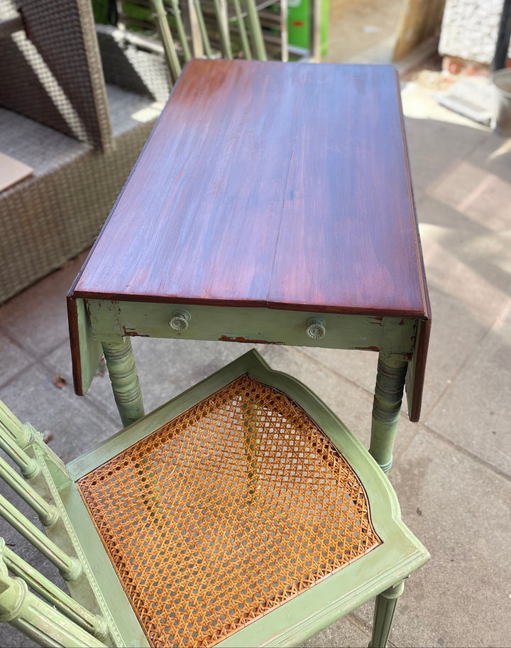 small rectangular wooden table with a dark reddish-brown top and a distressed sage green frame, shown in an outdoor patio setting next to a matching green chair with a woven cane seat.
Details