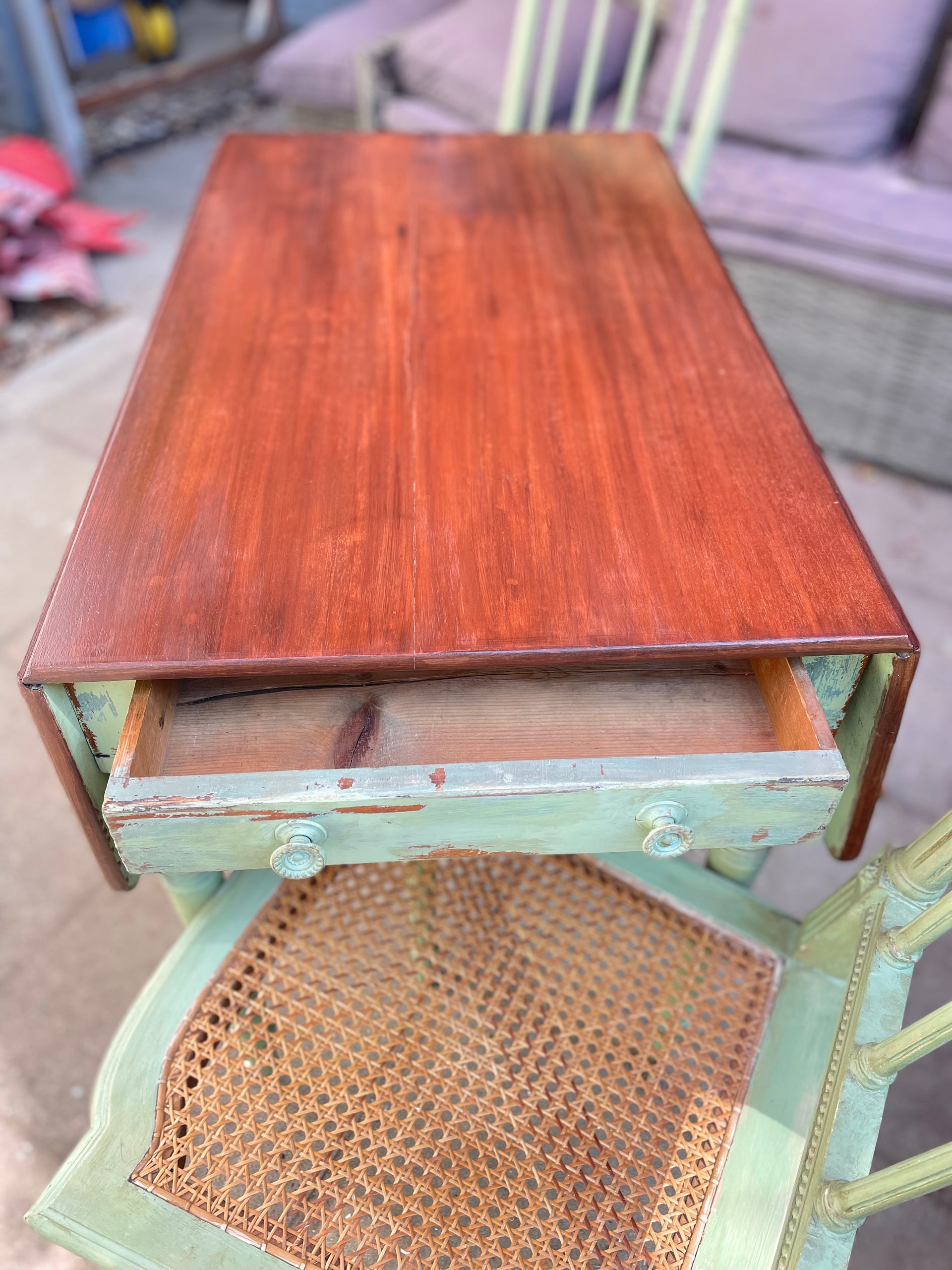 vintage wooden writing desk with a reddish-brown mahogany top and a distressed sage green painted frame, featuring an open drawer with a cane seat underneath.