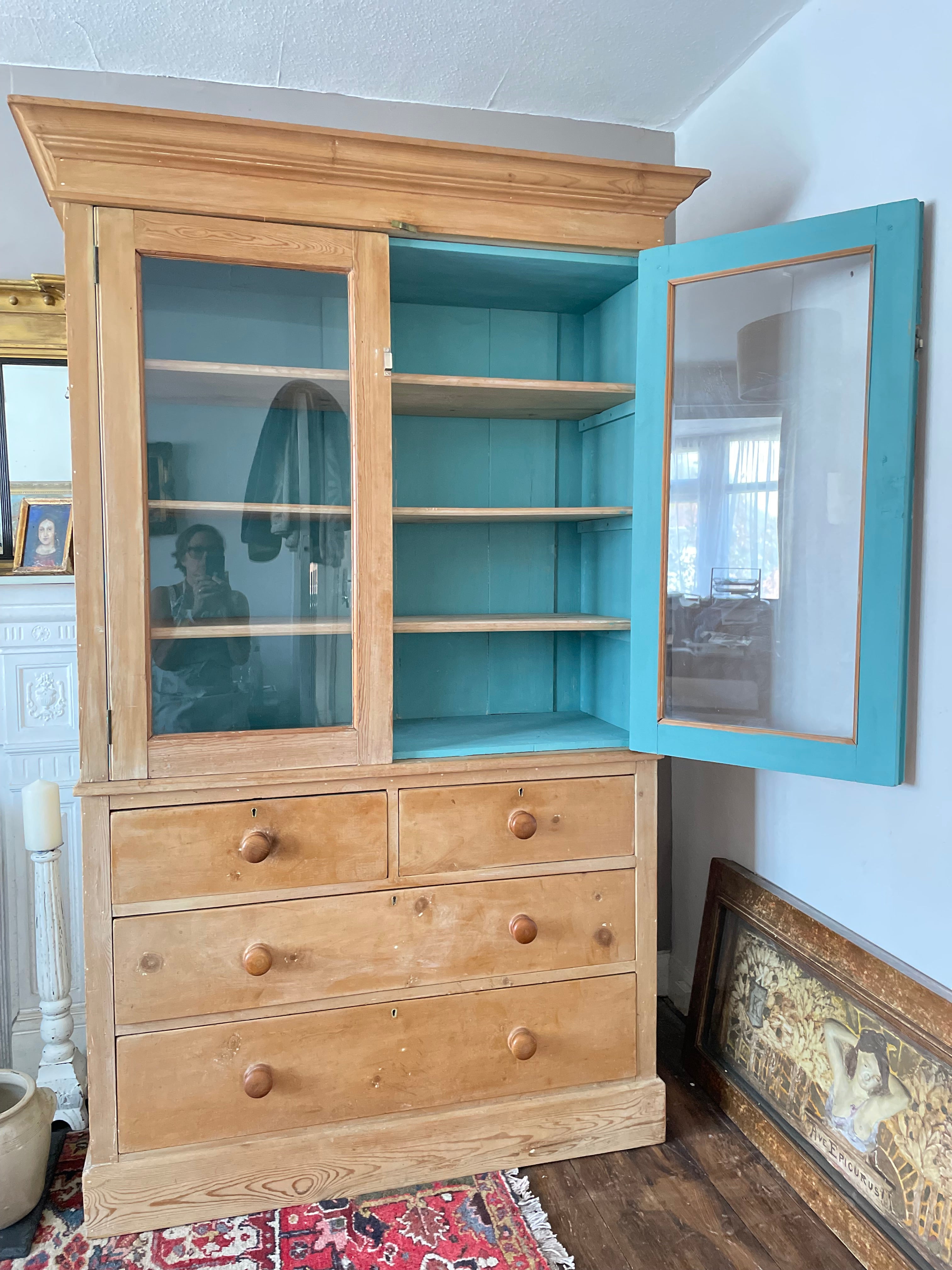 A vintage wooden display cabinet with a turquoise painted interior and glass-front doors, shown in a room setting with a patterned rug and framed artwork visible.