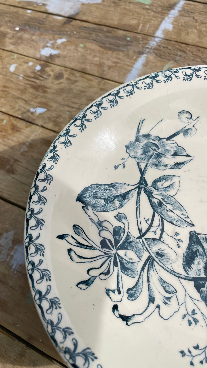 A close-up of a ceramic plate with a blue floral and leaf pattern on an off-white background, resting on a rustic wooden surface.